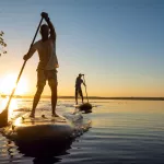 Men, friends sail on a SUP boards in a rays of rising sun. Stand up paddle boarding - awesome active recreation in nature. Backlight.